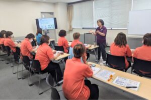 Instructor in a purple shirt lectures to adults in orange uniforms in a bright classroom with a presentation screen at the front.