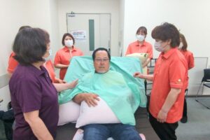 Man seated on a hospital bed wearing a green patient gown, surrounded by staff in masks in a clinical room